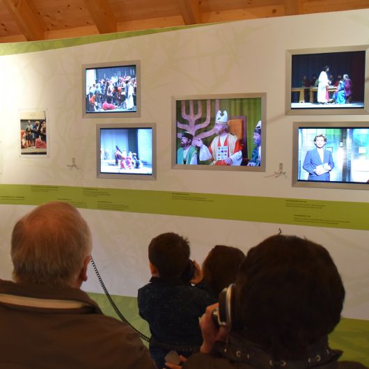 Besucher vor einer Wand im Museum, an der auf verschiedene Bildschirmen die Geschichte der Sömmersdorfer Passionsspiele gezeigt wird.