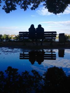 Ein Paar sitzt am 4. August 2015 auf einer Parkbank in Bonn und spricht miteinander. Ihre Silhouette spiegelt sich in einer Wasserpfütze.