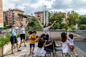 Kinder mit Mundschutz spielen mit Betreuerinnen auf einer Terrasse bei der Ferienbetreuung der Pfarrei San Bonaventura "Reise nach Jerusalem" am 18. Juni 2020 in Rom. Im Hintergrund: ein moderner Kirchturm mit Kreuz.