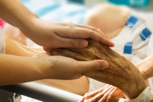 Zwei Hände halten die linke Hand eines alten Menschen. Die Szene ist an einem Krankenbett aufgenommen.