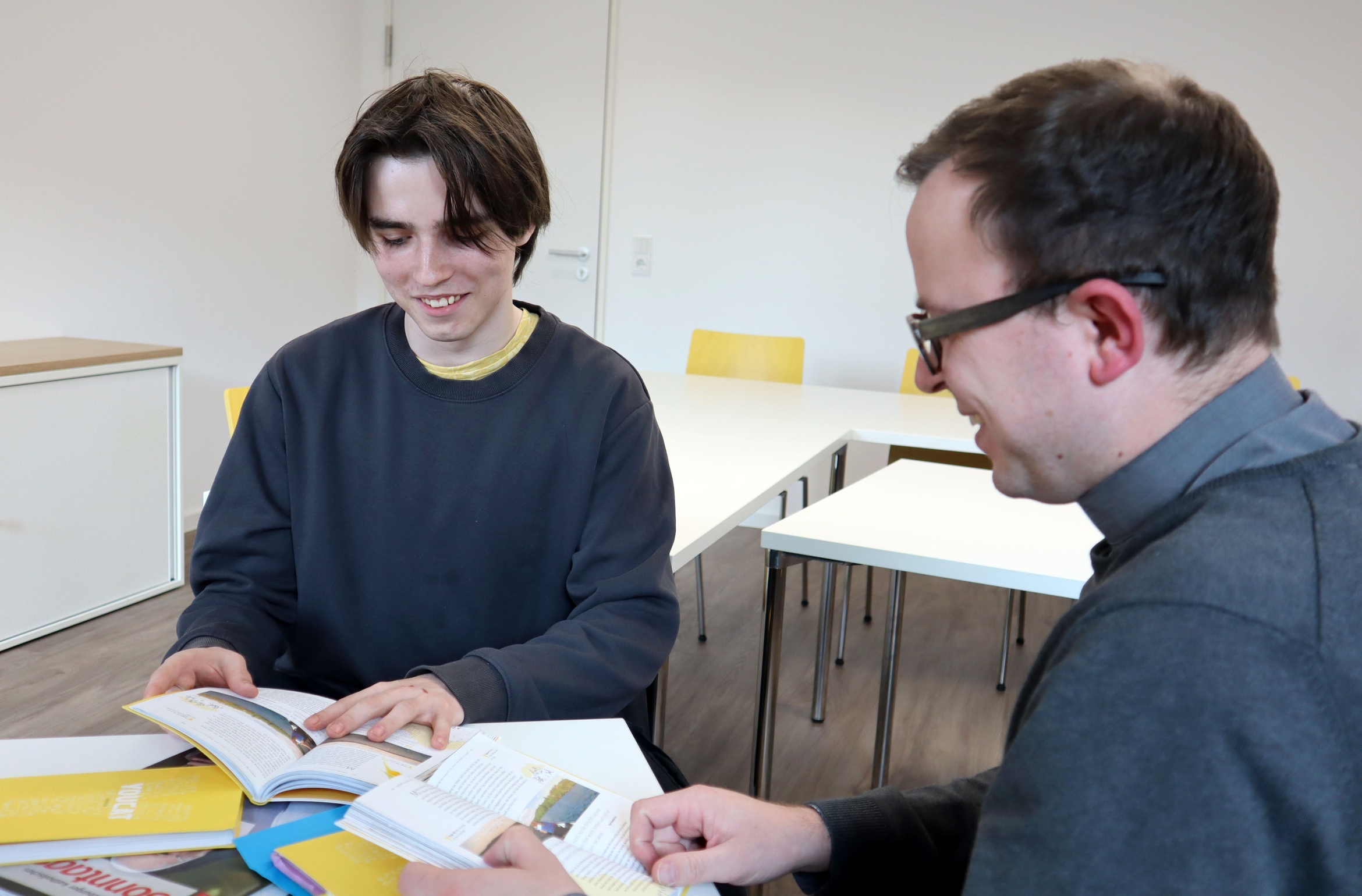 Noah Wirsching und Pfarrvikar Bertram Ziegler reden im Gemeindezentrum St. Andreas in Ochsenfurt. Auf dem Tisch vor ihnen liegen Bücher zur Taufvorbereitung.