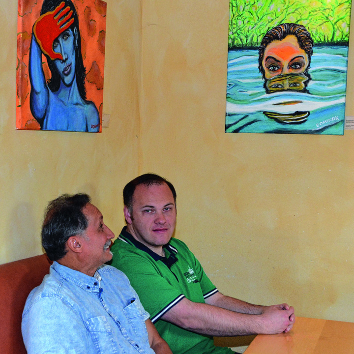 In einem Raum im Würzburger Café Perspektive sitzen auf einer Bank an einem Tisch Dominik Rösler (rechts) und José F. Sanchez. Über ihnen hängen zwei Bilder, unter anderem ihr eigenes mit dem Titel „Rothand“.