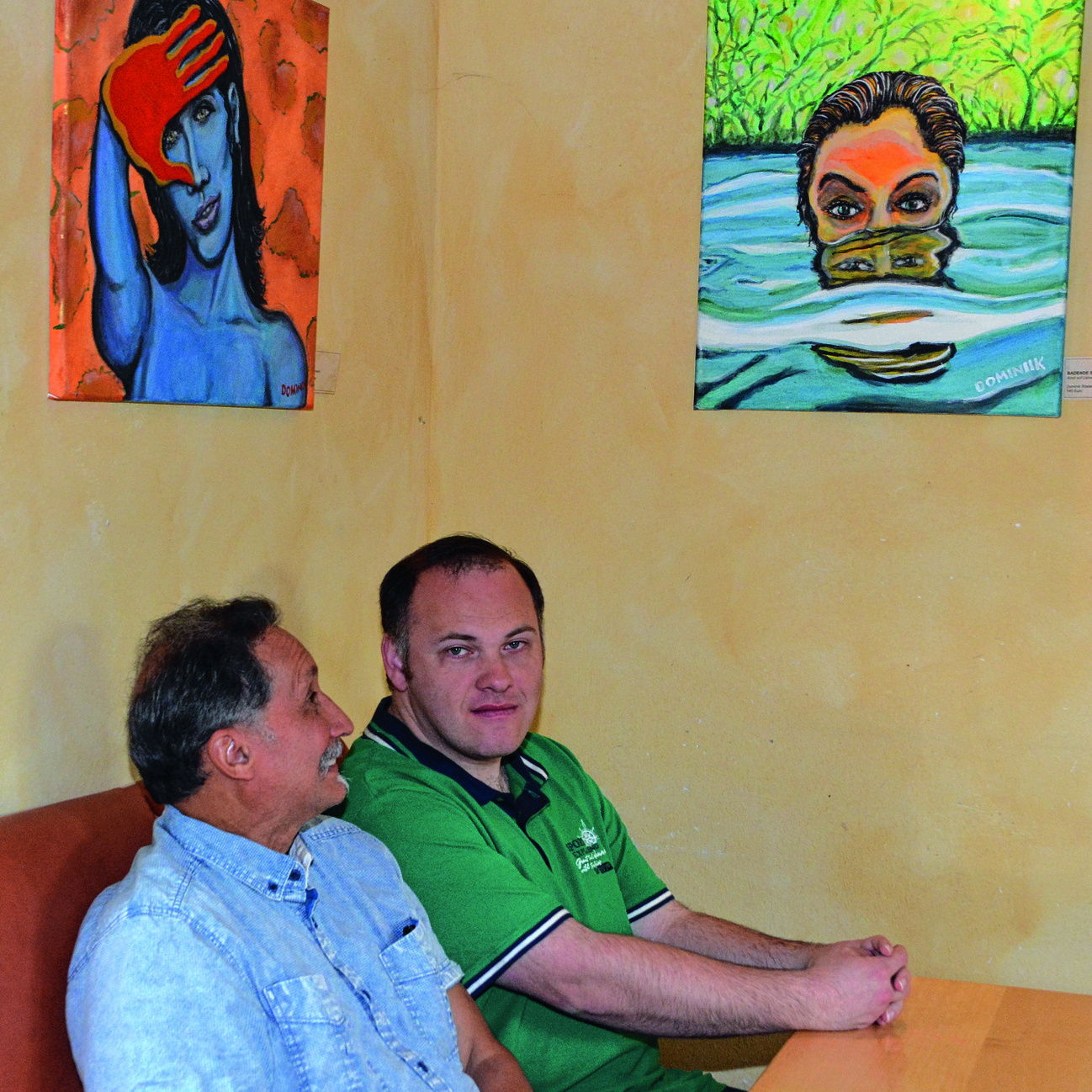 In einem Raum im Würzburger Café Perspektive sitzen auf einer Bank an einem Tisch Dominik Rösler (rechts) und José F. Sanchez. Über ihnen hängen zwei Bilder, unter anderem ihr eigenes mit dem Titel „Rothand“.