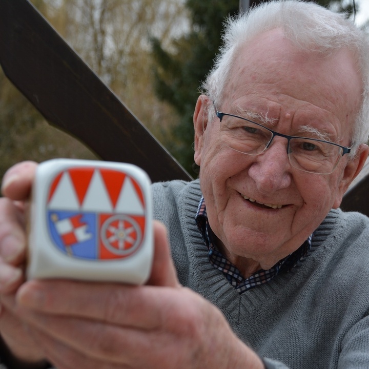 Wilhelm Wolpert hält den Frankenwürfel mit der Seite in die Kamera, die das unterfränkische Wappen zeigt. Dabei lächelt er.